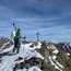 Skifahrer steht auf verschneitem Berggipfel mit Metallkreuz und Bergpanorama